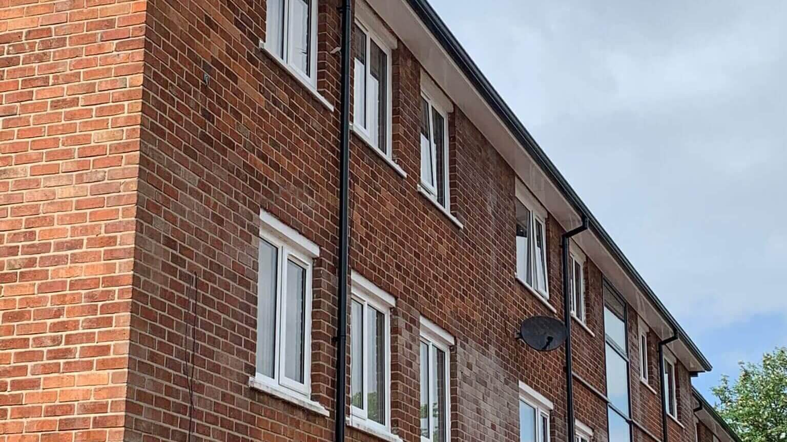 View from the ground up at a refurbished social housing block in Blackburn, showing newly fitted black uPVC gutters and downpipes against a cloudy blue sky, with green tree leaves visible to the bottom right of shot.