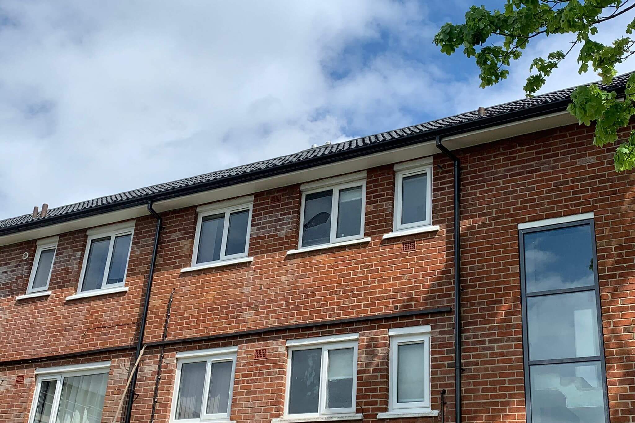 Ground-up view of a Blackburn housing block showing new black uPVC gutters and downpipes under a cloudy blue sky, with green tree branches on the right-hand side.