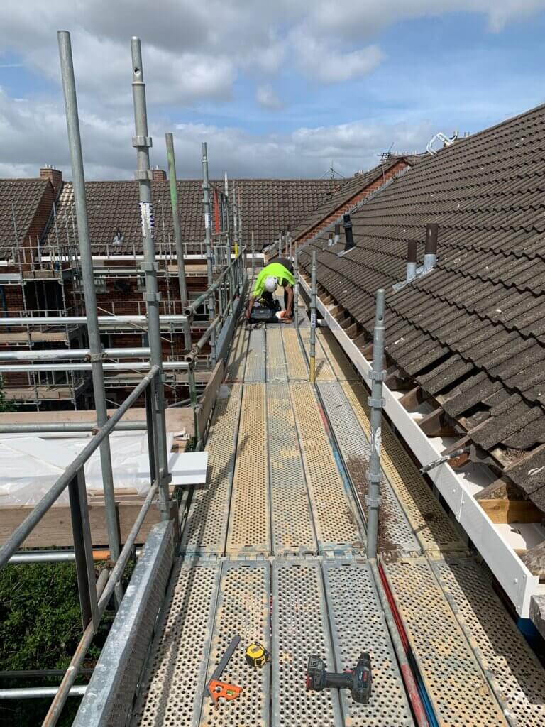 Installer in hi-vis and safety helmet working on scaffolding in Blackburn, bending over a bag of tools along a roofline installation run.
