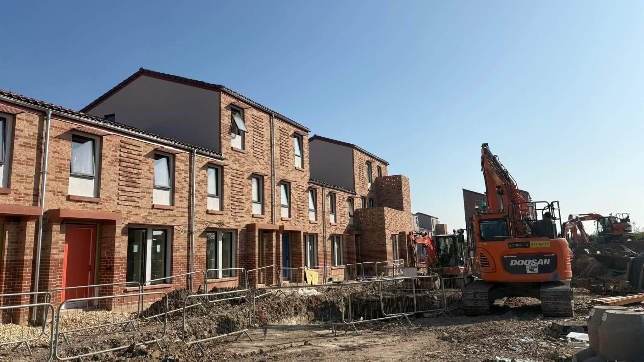 Row of new build houses with newly installed galvanised steel guttering and downpipes under clear blue sky, with three Doosan excavators and construction fencing visible at ground level