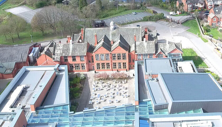 Aerial view of Sir John Deane's College, Nantwich, showing the Victorian red-brick main building alongside modern flat-roofed extensions, with the River Weaver and Nantwich town visible in the background.